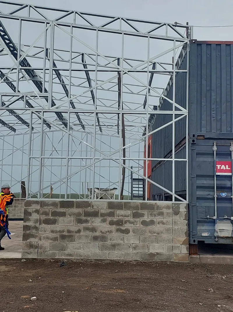 Steel roof and wall framing above a block wall beside shipping containers.